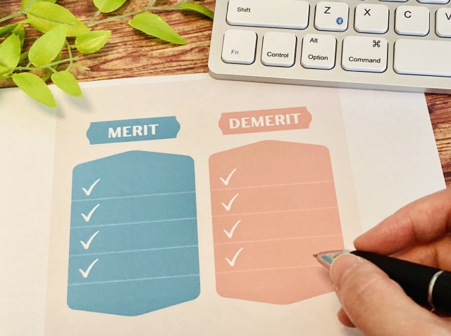 机の上にPCと「メリット」「デメリット」と書かれた紙があり、ペンを持つ人の指がうつっている